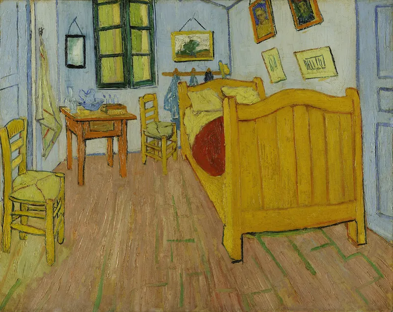De slaapkamer van Vincent van Gogh: een kamer met blauwe muren, een houten bed met rode deken en twee kussens, twee stoelen, een tafeltje met waskom en schilderijtjes aan de wand.